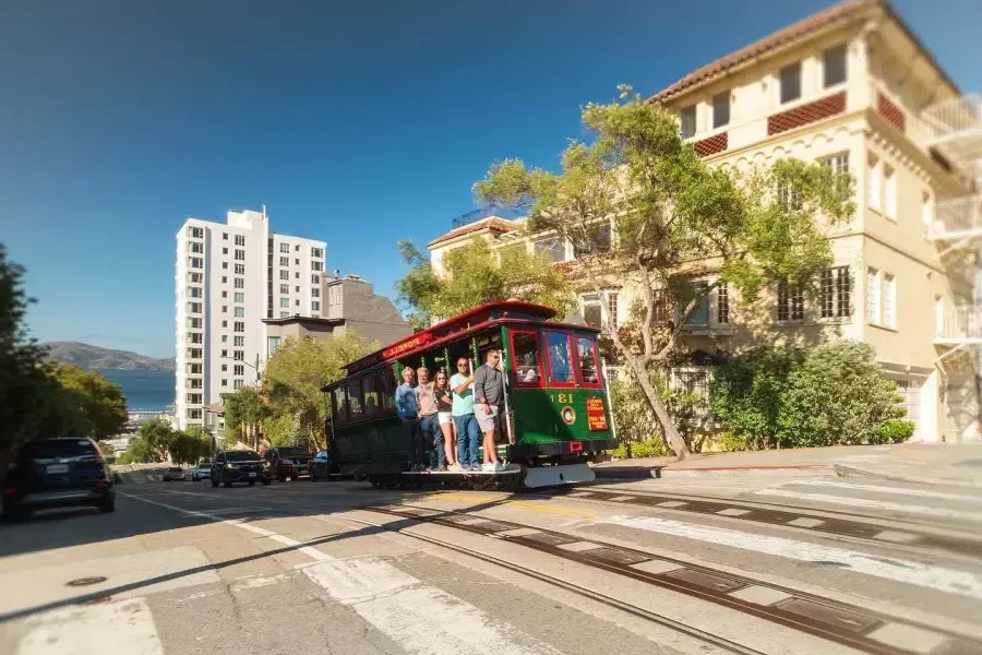 Powell and Hyde cable car going up the street with smiling people inside on a sunny day.